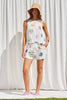 Woman wearing a sleeveless top and shorts with floral and palm tree patterns against a white curtain.